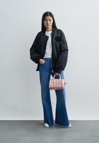 Mujer con chaqueta bomber negra, camiseta blanca, jeans de pierna ancha azules, zapatillas blancas, sosteniendo un bolso de mano de estampado rosa, de pie sobre un suelo y una pared grises.