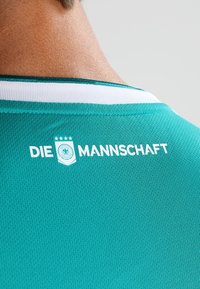 Zadní strana športového dresu v modrozelené barvě s bílým obšitím límce, obsahující text "DIE MANNSCHAFT" a čtyřhvězdičkový emblém s logem německého orla.