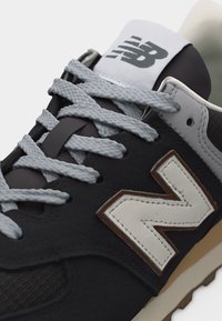 Ασπρόμαυρο αθλητικό παπούτσι New Balance με γκρι κορδόνια, λευκό λογότυπο "N" με καφέ περίγραμμα και μπεζ σόλα ορατή στην κάτω άκρη.