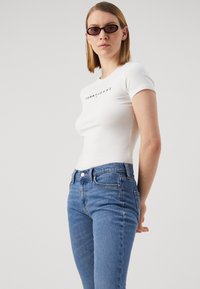 T-shirt bianca aderente con logo nero "TOMMY JEANS", abbinata a jeans in denim blu. Il modello ha i capelli corti e indossa occhiali da sole ovali piccoli.