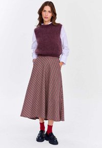 Žena oblečená v fialovej bezrukávovej svetríku cez bielu košeľu, s midi sukňou s červenými a čiernymi kockami, červenými ponožkami a čiernymi mokasínami.