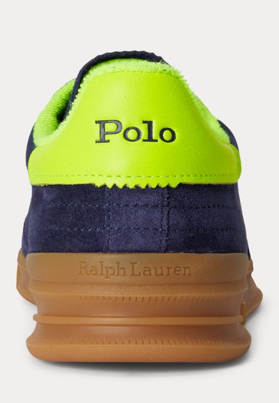 Ναυτικό σουέντ αθλητικό παπούτσι με ταμπέλα φτέρνας σε νέον πράσινο χρώμα με την επιγραφή "Polo" και σόλα από καουτσούκ χρώματος τσίχλας με ανάγλυφη επιγραφή "Ralph Lauren", εμφανιζόμενο από πίσω.