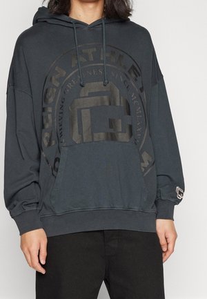 Personne portant un sweat à capuche sombre oversize avec un grand graphisme circulaire et du texte, une poche avant et des manches longues, associé à un pantalon noir.