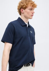 Νέος άνδρας με κατσαρά κόκκινα μαλλιά, φοράει μπλε σκούρο πόλο πουκάμισο Calvin Klein και ανοιχτόχρωμο παντελόνι, στέκεται μπροστά σε ένα απλό φόντο.