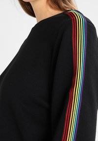 Czarny sweter z okrągłym dekoltem i długimi rękawami, zdobiony pionowymi tęczowymi paskami wzdłuż zewnętrznego szwu na ramieniu.