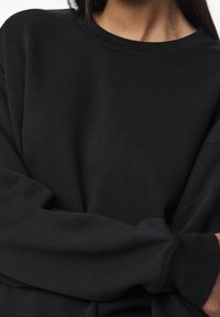 Černá dlouhá mikina s dlouhým rukávem vyrobená z měkkého materiálu. Disponuje kulatým výstřihem a mírně oversized střihem, s viditelnými švy na rukávech.