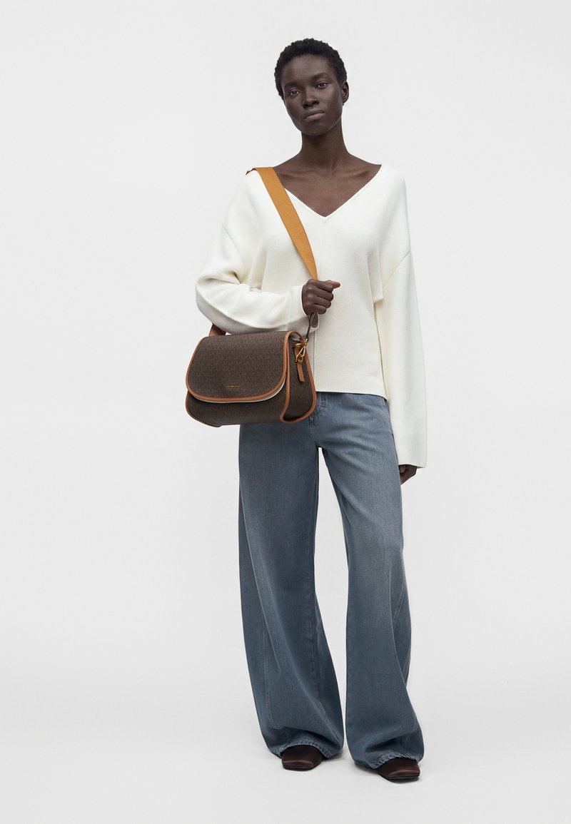 Femme portant un pull blanc à col en V et un jean bleu à jambes larges, tenant un sac bandoulière marron avec une sangle orange sur un fond uni.
