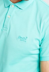 Polo turquoise en tissu texturé, avec une patte de boutonnage à deux boutons et un logo brodé de la même couleur sur la poitrine.