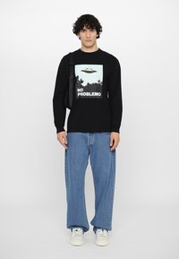 Black long-sleeve shirt with a graphic of a UFO and "NO PROBLEMO" text, paired with light blue wide-leg jeans and white sneakers.