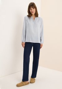 Femme se tenant debout, portant un chemisier à rayures bleu et blanc à manches longues, un jean droit bleu foncé et des chaussures sans lacets beiges.