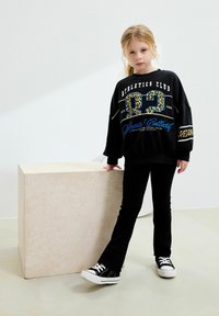 Svart sweatshirt med leopardmönstrad siffra "82" och vit text, i kombination med svarta byxor och svarta sneakers med vita sulor, stående vid en stenliknande kub.