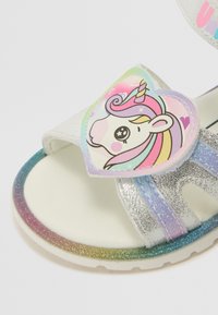 Weiße Sandalen mit glitzerndem Finish, featuring ein farbenfrohes Einhorn-Design auf einem herzförmigen Patch und einer Regenbogenverlaufsohle.