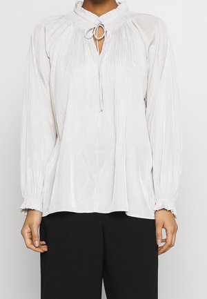 Person iført en hvid bluse med lange ærmer med plisserede detaljer, flæsekant, bindelukning ved halsen og sorte bukser.