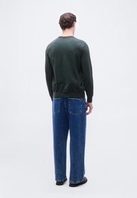 Άνδρας που στέκεται πλάτη, φοράει σκούρο πράσινο πουλόβερ, φαρδιά μπλε τζιν και μαύρα παπούτσια, πάνω σε ανοιχτόχρωμο απλό φόντο.