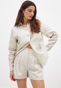 Vaalea beige pellavapaita, jossa on nappikaulus ja yksi rintatasku, yhdistettynä korkeavyötäröisiin shortseihin, joissa on laskokset.