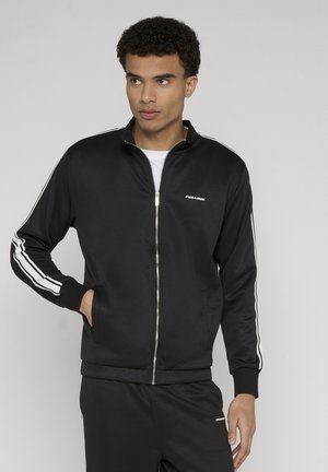 Joven con chándal negro de cremallera completa con rayas blancas en las mangas y pantalones, de pie con una mano en el bolsillo, mirando de lado.