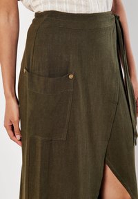 Olivenfarbene Wickelrok aus leichtem Stoff. Verfügt über eine große Tasche mit zwei Metallknöpfen und einem Bindebandverschluss in der Taille.