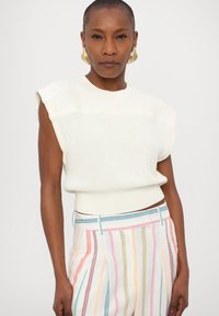 Bela pletena brezrokavna majica z rebrasto teksturo, v kombinaciji z večbarvnimi prugastimi hlačami z navpičnimi črtami in poudarjenim pasom.