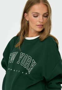 Sudadera verde oscuro con gráfico "NEW YORK" en blanco. Presenta un cuello redondo y un ajuste holgado, acentuada por un cuello interior blanco.