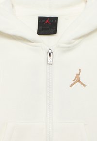 Κρεμ φούτερ με φερμουάρ, κατασκευασμένο από απαλό ύφασμα, με τσέπη μπροστά, ασημί φερμουάρ και χρυσό κεντημένο λογότυπο Jumpman στην αριστερή πλευρά.