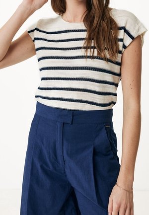 Femme portant un haut blanc sans manches à rayures horizontales bleu marine et un pantalon bleu marine taille haute, main près du visage.