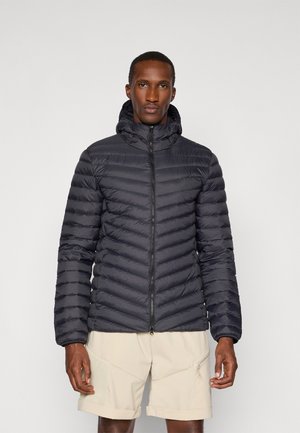 Zwarte pufferjas met een ritssluiting, capuchon en horizontaal gestikte design. Gedragen over lichtbeige shorts. Lichtgewicht en geïsoleerd materiaal.
