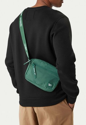 Mann trägt schwarzen Pullover und beige Hose mit einer grünen Reebok Umhängetasche, die über Rücken und Schulter hängt.