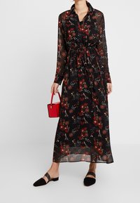 Langes schwarzes Kleid mit rotem Blumenmuster, halbtransparentem Stoff, Knopfleiste vorne, geraffter Taille und langen Ärmeln. Ergänzt durch eine kleine rote Handtasche.