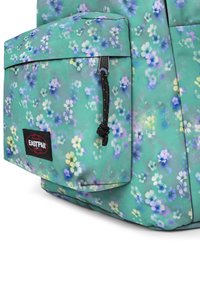 Mochila verde con estampado floral que presenta un bolsillo frontal con una cremallera negra y logo de Eastpak. El material parece suave con una textura mate.