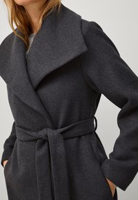 Cappotto in lana grigio scuro con ampio risvolto, vita cintata e tasche laterali, caratterizzato da un tessuto testurizzato e una silhouette fluida.