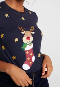 Marinblå t-shirt med en ren med röd nos, grön scarf och en julstrumpa, prydd med glittrande gyllene stjärnor.