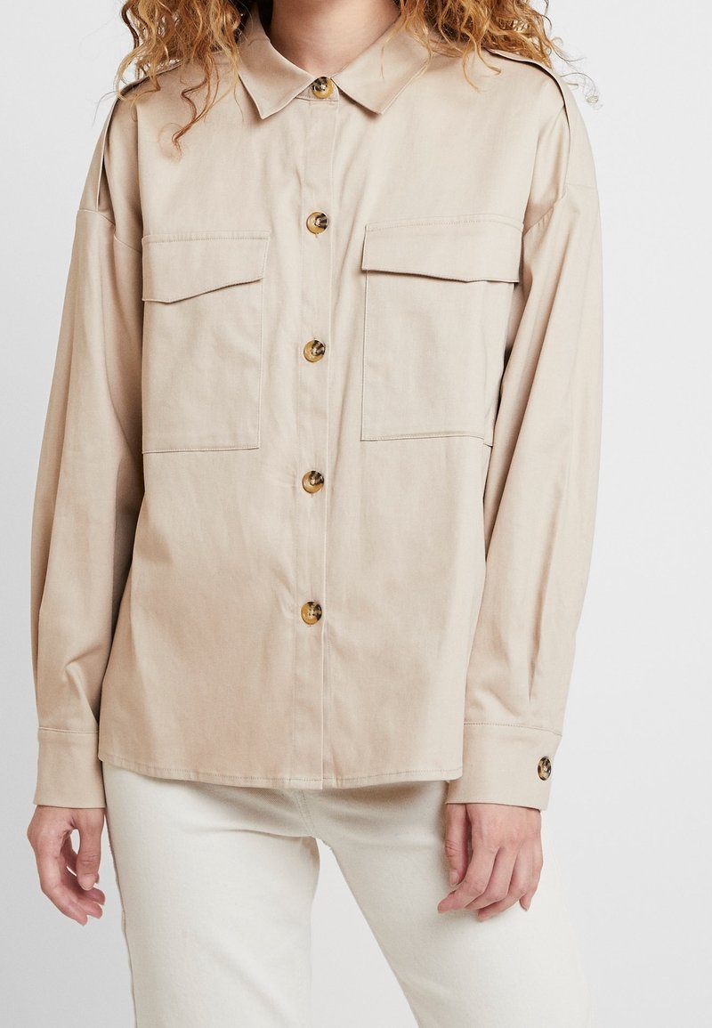 Chemise beige à boutons avec col, deux poches poitrine et manches longues. Dotée de boutons dorés et d'une coupe décontractée.