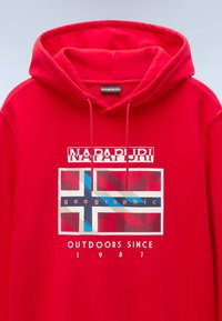 Sudadera roja con capucha que presenta un diseño gráfico con un motivo de bandera, el nombre de la marca "NAPAPURI" y el texto "AL AIRE LIBRE DESDE 1987".