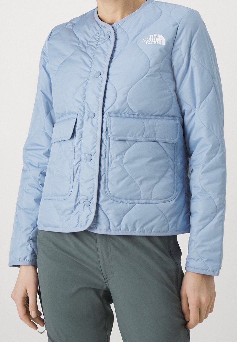Jasnoniebieska pikowana kurtka z zapięciem na guziki, dwoma przednimi kieszeniami oraz logo North Face na lewej stronie klatki piersiowej.