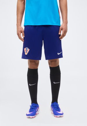 Atlet masculin purtând pantaloni scurți de fotbal Croația albaștri, cu șosete negre Nike și ghete albastre Nike, stând în fața unui fundal simplu.