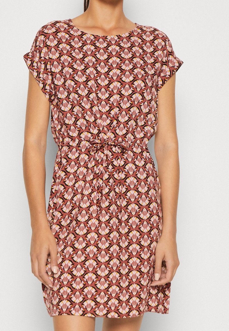 Femme portant une robe à manches courtes, longueur genou, avec un motif floral géométrique rose et marron et une ceinture nouée à la taille.