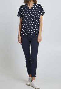 Navy blue short-sleeve top with white polka dots, V-neck design, and a relaxed fit, paired with dark pants and white sneakers.