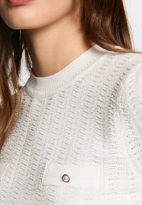 Biały sweter dzianinowy z teksturowanym wzorem, prążkowanym dekoltem oraz dwiema kieszeniami z guzikami przypominającymi perły.
