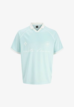 Teenager iført solbriller, lyseblå skjorte, falmede vidde jeans og tanfarvede støvler, stående og smilende mod en ensfarvet hvid baggrund.