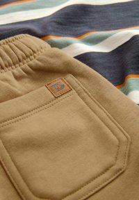 Khaki sweatpants med elastisk talje og en baglomme med en syet lysebrun label. Baggrunden inkluderer et stribet stof i afdæmpede farver.