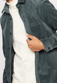 Primo piano di una persona con indosso un maglione bianco a collo alto sotto una giacca in velluto a coste verde scuro sbottonata, con la mano che tiene aperta la giacca.