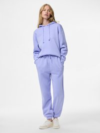 Nő áll, lila kapucnis pulóvert és hozzáillő melegítőnadrágot visel fehér sportcipővel, kezei zsebre dugva, tiszta fehér háttér előtt.