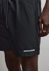 Schwarze Shorts mit Kordelzug, getragen von einer dunkelhäutigen Person, die einen silbernen Ring an der Hand und das "PEGADOR"-Logo am Bein zeigt.
