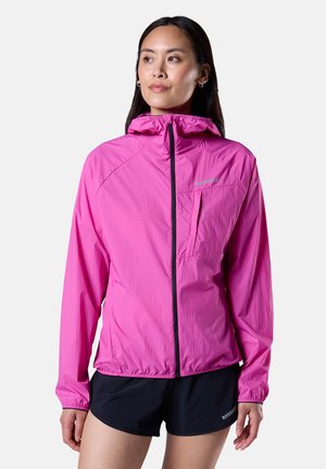 Nainen pukeutuneena kirkkaanpunaiseen kevyttoppatakkiin, jossa on etuvetoketju ja huppu, yhdistettynä mustiin urheilushortseihin, seisomassa yksinkertaisen taustan edessä.