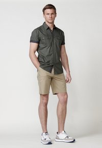 Camisa de manga corta verde oliva con dos bolsillos frontales, combinada con pantalones cortos beige y zapatillas deportivas blancas con detalles en gris y azul marino.
