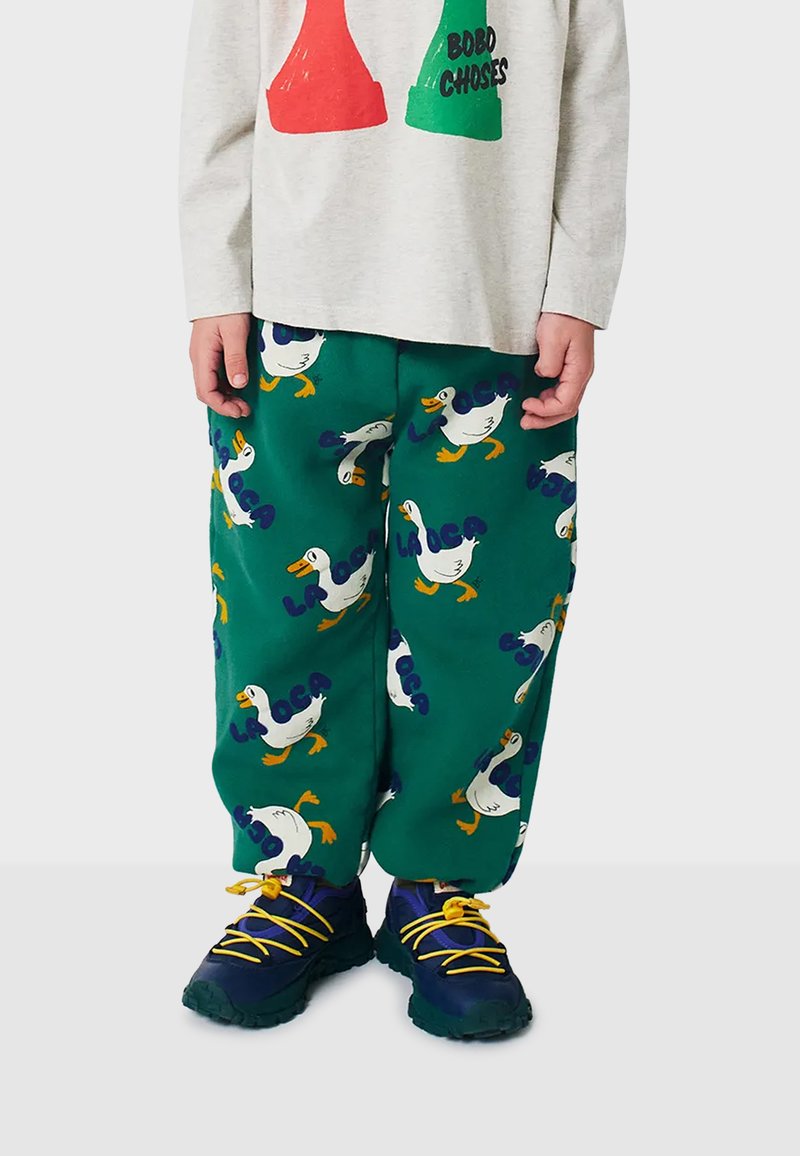 Green pants with white goose patterns and blue text, paired with navy sneakers featuring yellow laces. Soft fabric, loose fit.