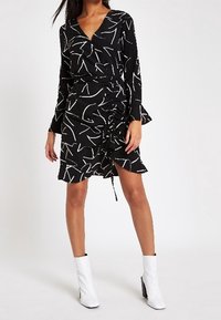 Svart klänning med vita abstrakta mönster, med långa ärmar, omlottdesign, rynkad detalj på sidan och volangkant. Bärs med vita ankelstövlar.