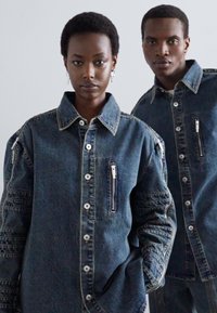 Jachetele din denim cu o spalare inchisa au siluete oversized, inchideri cu nasturi, buzunare cu fermoar si maneci texturate cu detalii cusute.