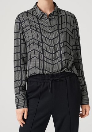 Nainen, joka on pukeutunut mustavalkoiseen, geometrisella kuviolla koristeltuun pitkähihaiseen paitaan, joka on työnnetty mustien nyöriviritteisten housujen sisään, seisoo yksiväristä taustaa vasten.