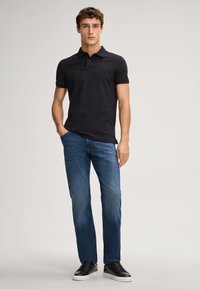 Zwarte polo-shirt met korte mouwen en een knoopsluiting, gecombineerd met blauwe spijkerbroek en zwarte sneakers op een neutrale achtergrond.